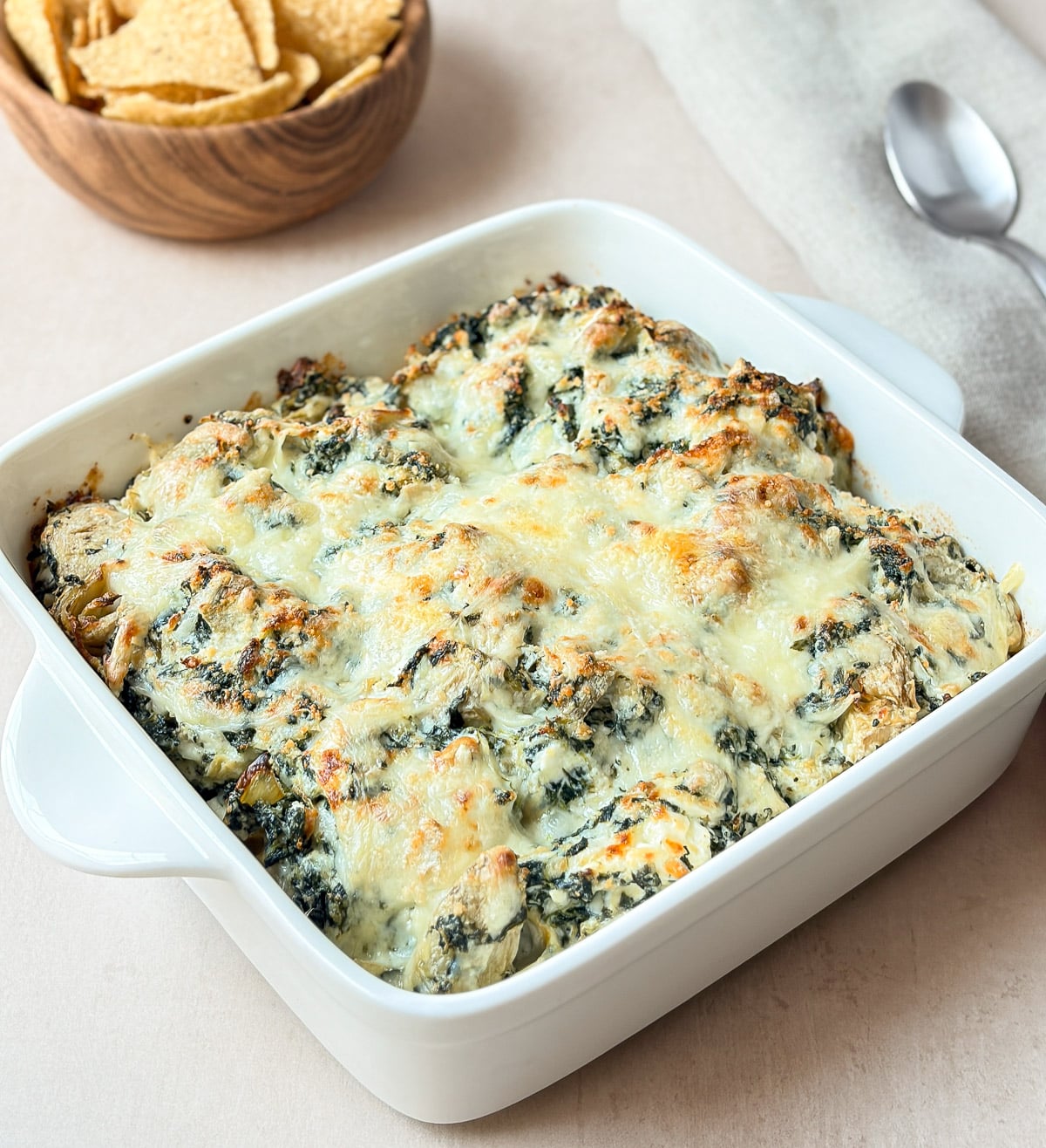 Creamy spinach artichoke dip baked in a white square dish with a golden cheesy top, served with tortilla chips.