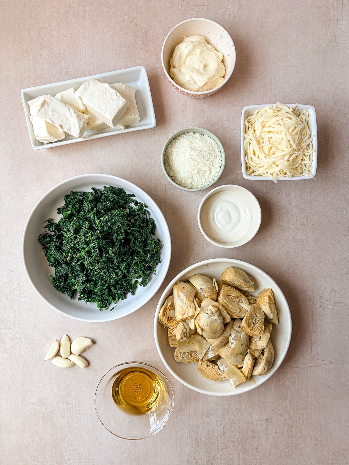 Spinach artichoke dip ingredients arranged on a tan surface, including spinach, artichokes, cream cheese, mozzarella, Pecorino Romano, sour cream, mayonnaise, garlic, and olive oil.