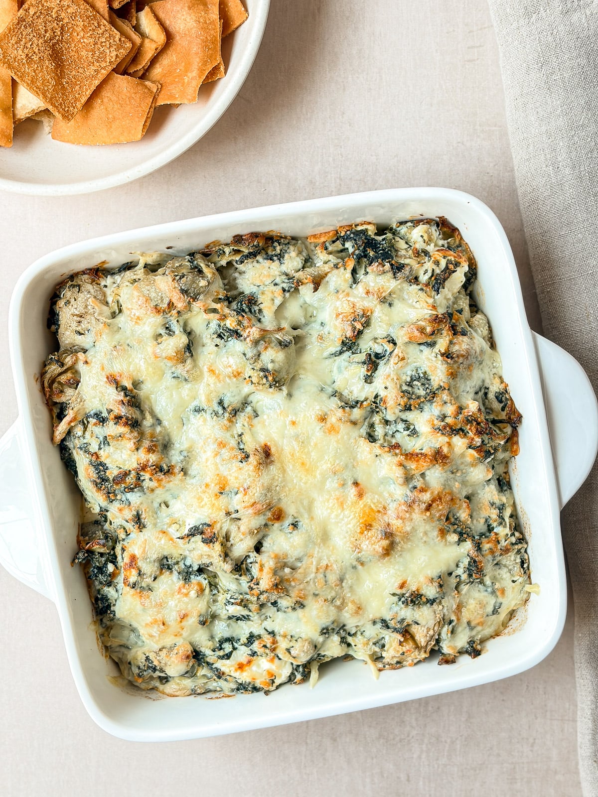 Overhead view of baked spinach artichoke dip with a golden cheesy top in a white square dish beside pita chips.