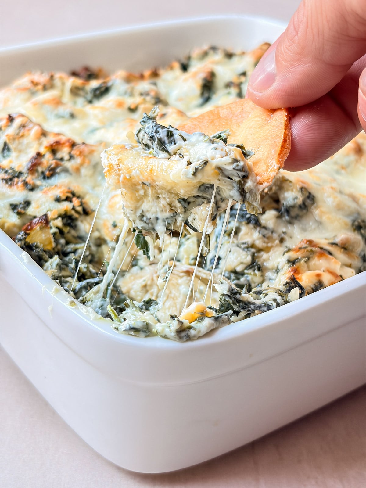 A tortilla chip lifting a cheesy scoop of spinach artichoke dip with melted cheese stretching from the baking dish.