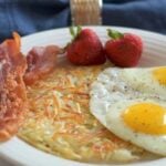 White plate with crispy golden shredded hashbrowns, 2 eggs sunny side up, 2 slices bacon, 2 strawberries with blue napkin and glass of milk